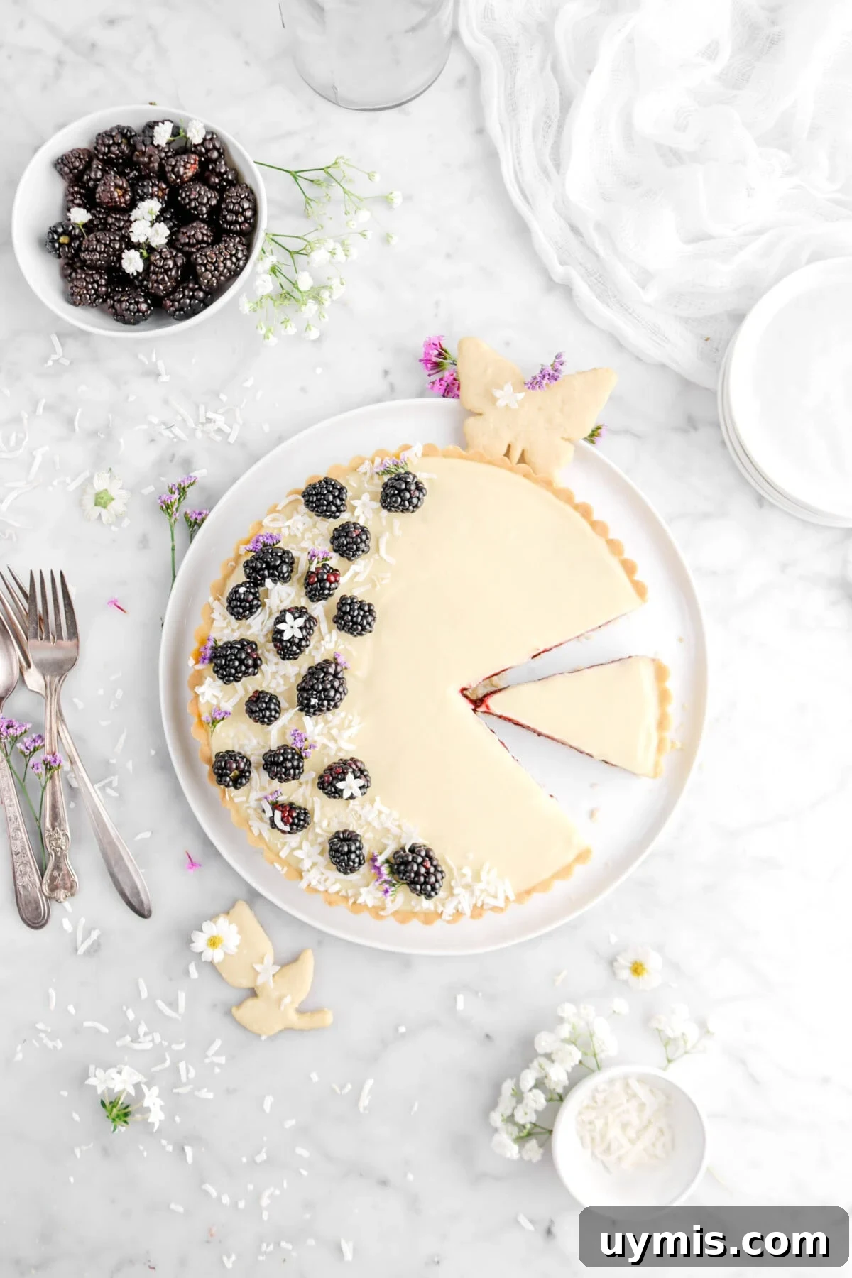 overhead shot of cut coconut blackberry with slice beside on white plate, with butterfly cookie and tulip cookie, fresh blackberries, forks, and purple flowers