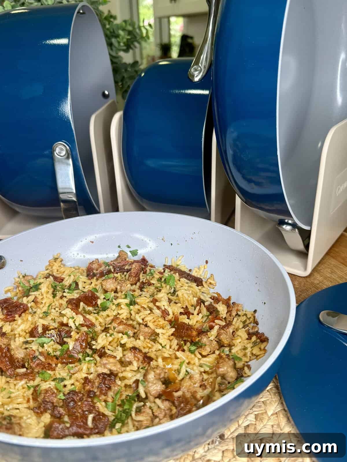 Why I Switched to Caraway Cookware: One-Pan Dinner for Two 2 A close-up of a pan filled with cooked rice, ground meat, and herbs sits on a woven placemat, next to a set of blue and gray nonstick cookware arranged upright in a kitchen setting.