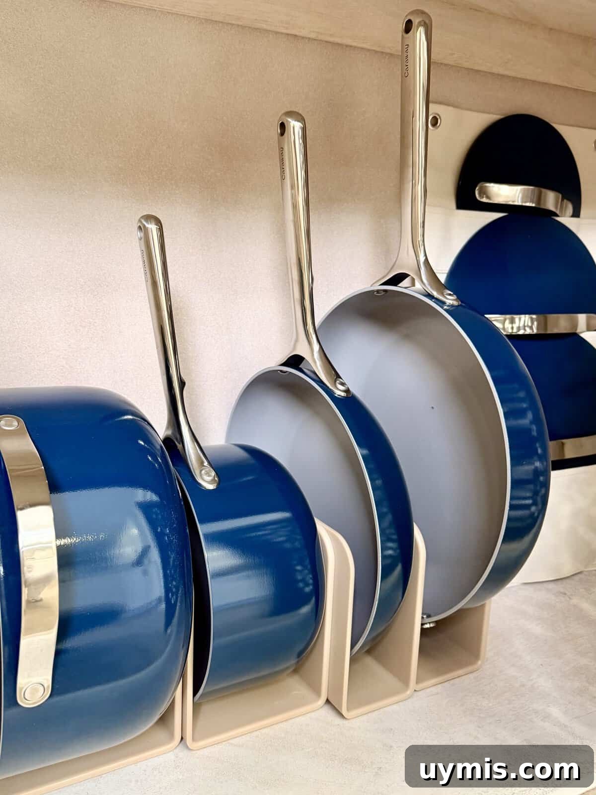 Why I Switched to Caraway Cookware: One-Pan Dinner for Two 3 Three blue and silver frying pans are neatly organized upright in a beige rack. Inspired by Why I switched to Caraway Cookware, their matching blue lids stand vertically nearby on a light-colored shelf, ready for an easy one-pan dinner for two.