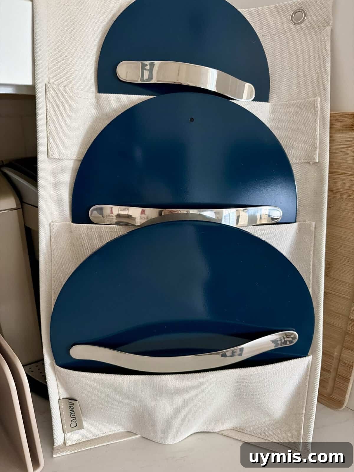 Why I Switched to Caraway Cookware: One-Pan Dinner for Two 5 Three blue pot lids with silver handles are stored vertically in a white fabric organizer, which is standing on a kitchen counter next to a wooden cutting board and other kitchen items.