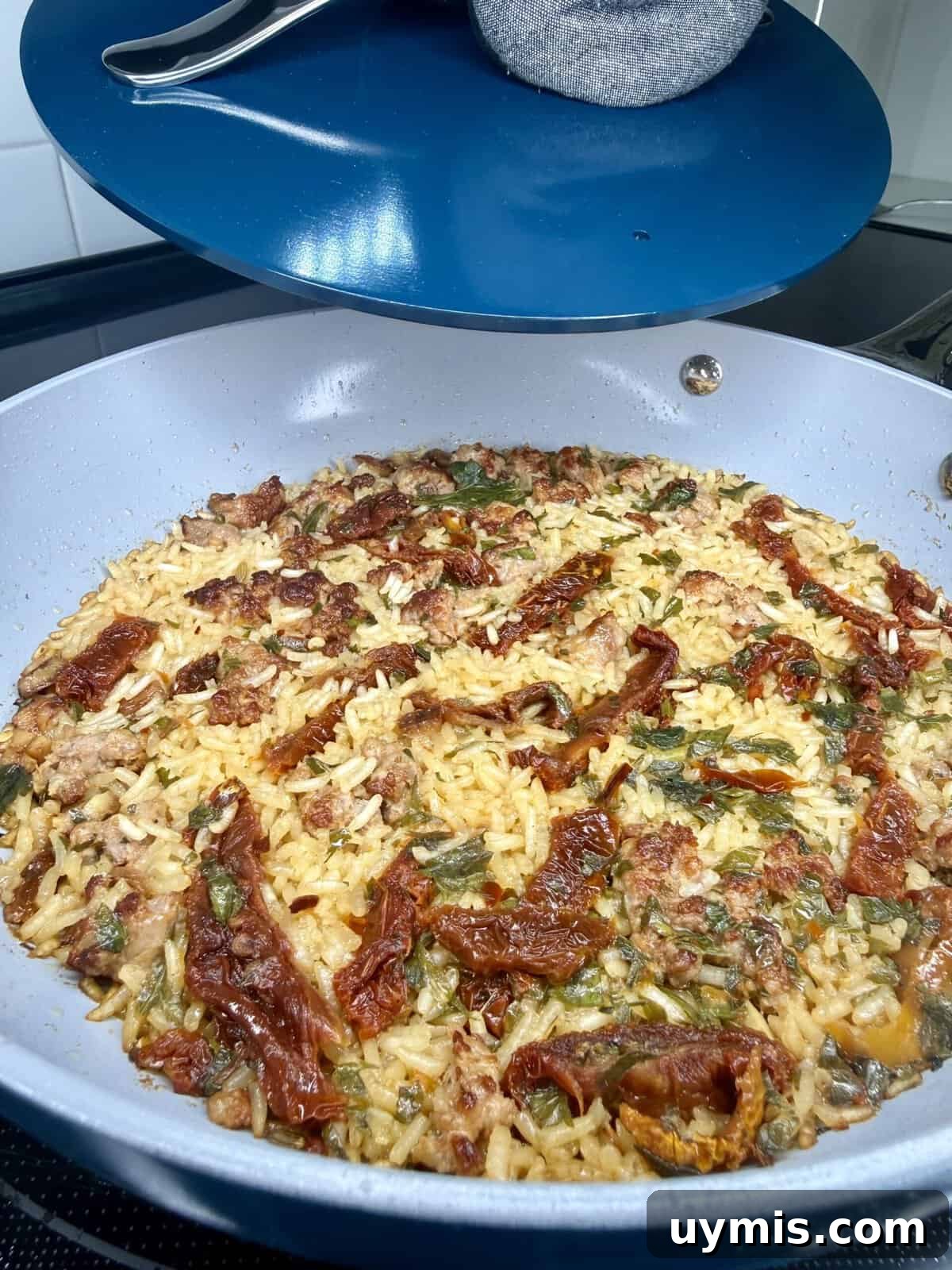 Why I Switched to Caraway Cookware: One-Pan Dinner for Two 6 A close-up of a pan filled with cooked rice, sun-dried tomatoes, green herbs, and ground meat, sitting on a stove with a blue lid partially covering the pan.