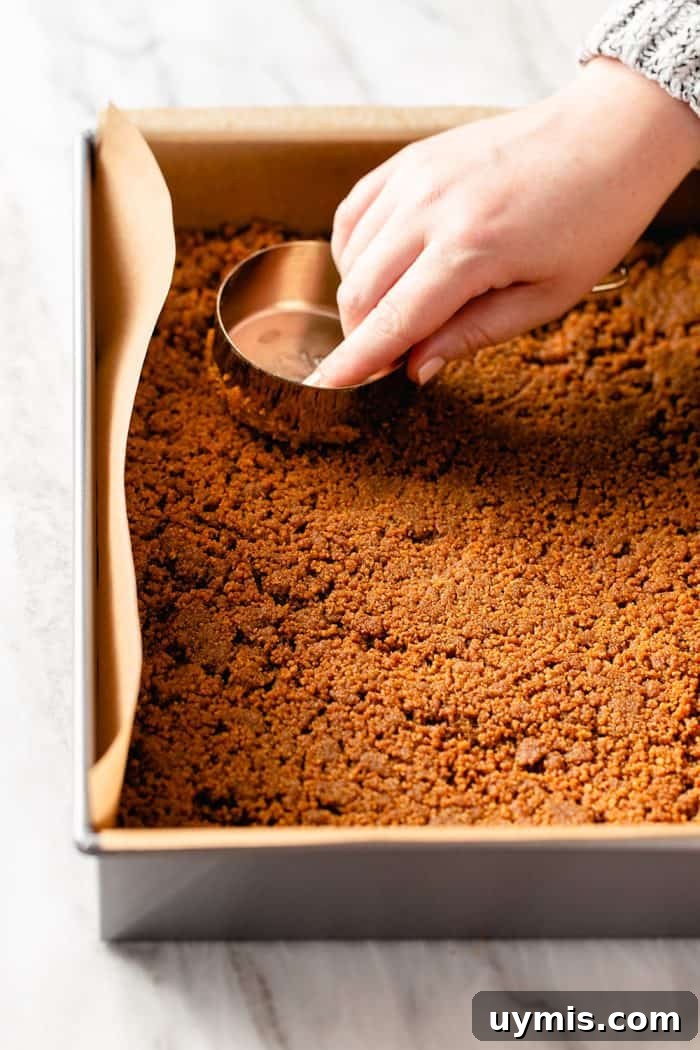 Hand using a measuring cup to press a cookie crust into a parchment-lined pan
