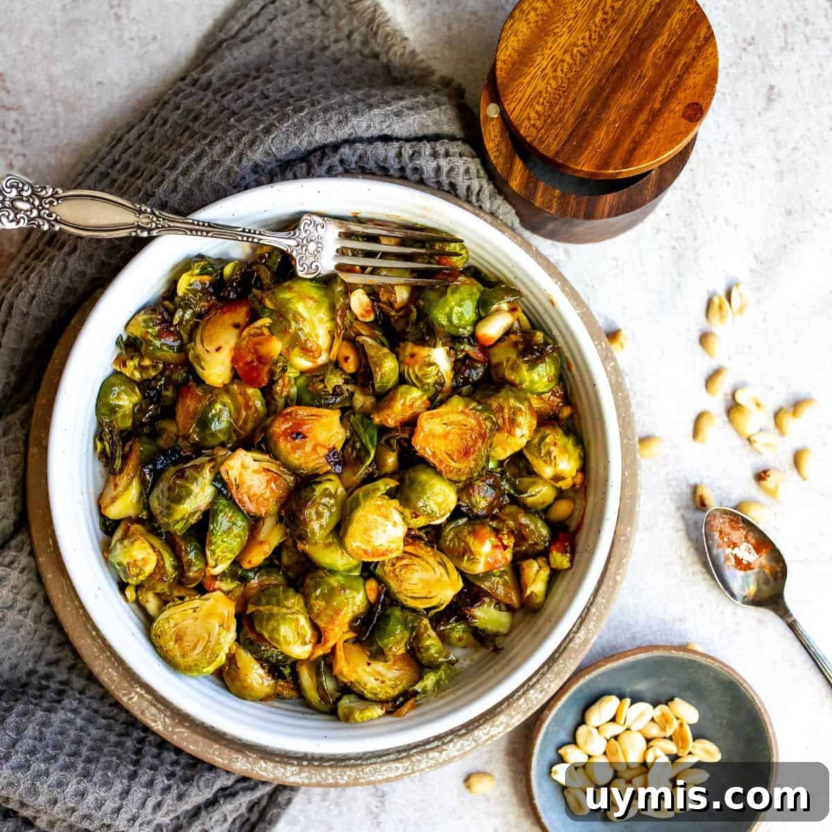Crispy Honey Sriracha Brussels Sprouts 2 White bowl filled with honey sriracha brussels sprouts and sauce with a silver spoon and peanuts on the side.