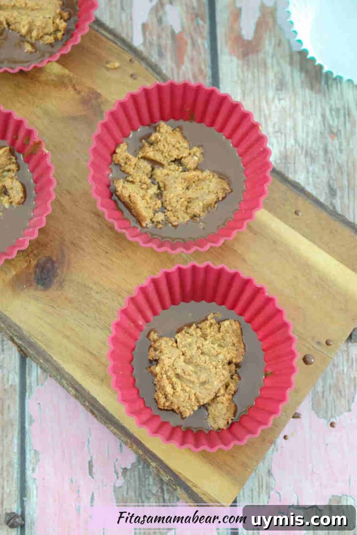 High-Protein Almond Butter Dark Chocolate Cups 6 Res silicone muffin cups with melted chocolate and almond butter on a wooden cutting board.