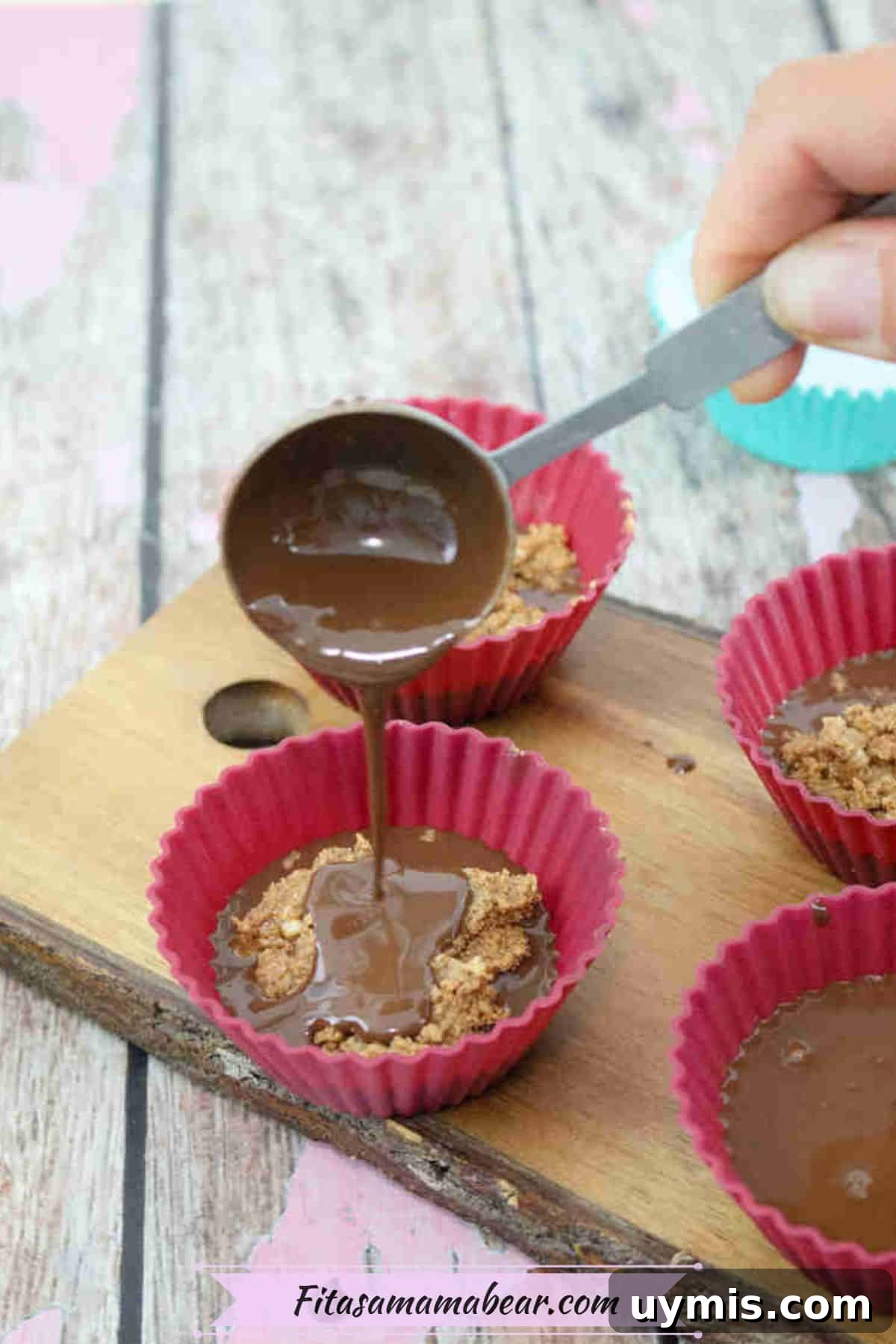 High-Protein Almond Butter Dark Chocolate Cups 7 Red silicone molds on a wooden cutting board with melted chocolate being poured over almond butter.