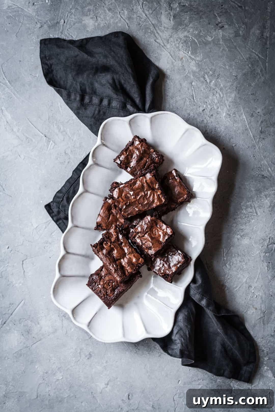 Chocolate Brownies with Port-Soaked Cherries 8 Top view of white fluted platter with cut squares of brownies with port soaked cherries on a grey background with a dark grey linen napkin