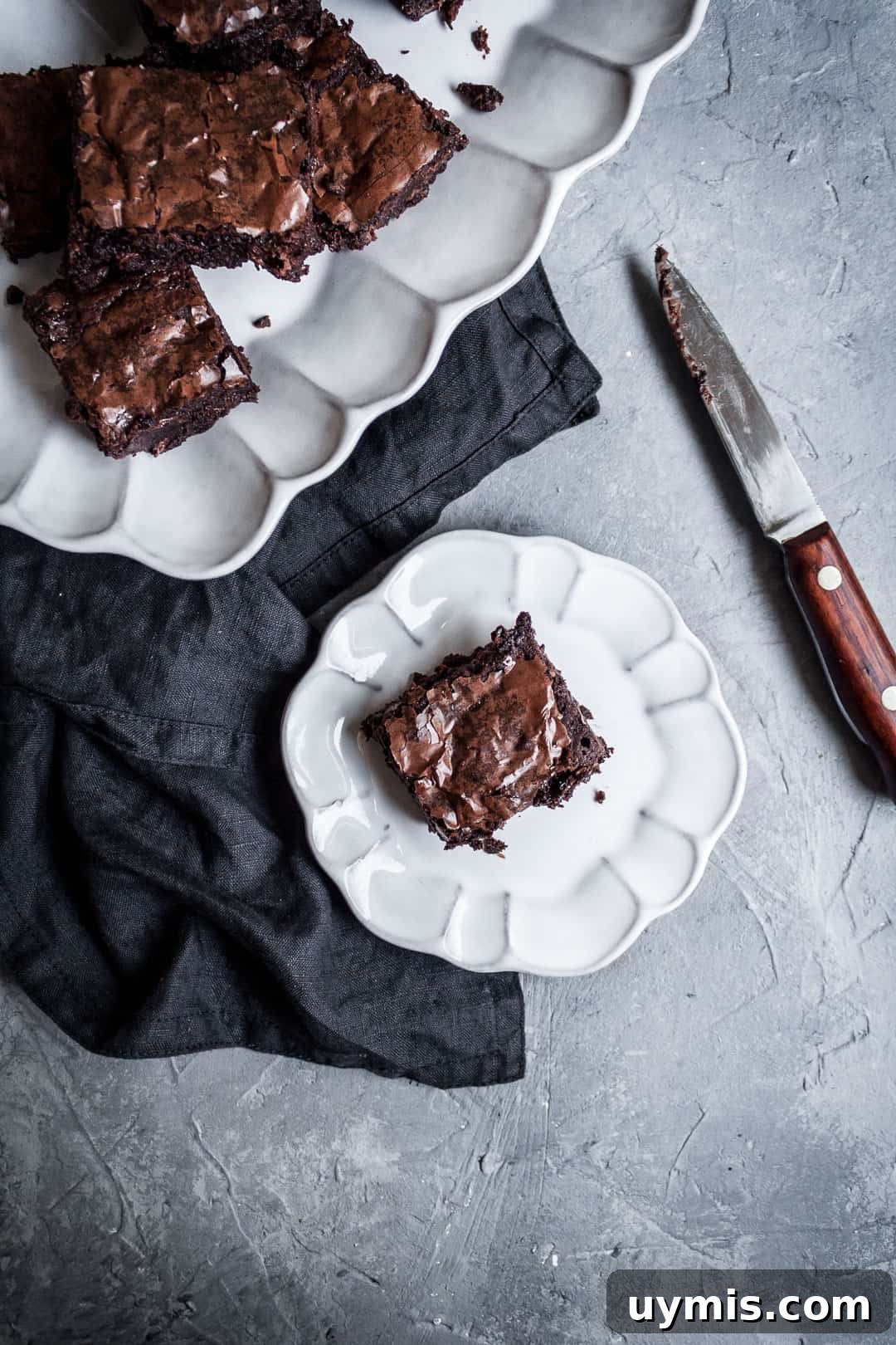 Chocolate Brownies with Port-Soaked Cherries 6 White plate with a cut square of brownies with port soaked cherries, a dark grey linen napkin and a wooden handled knife all on a grey background