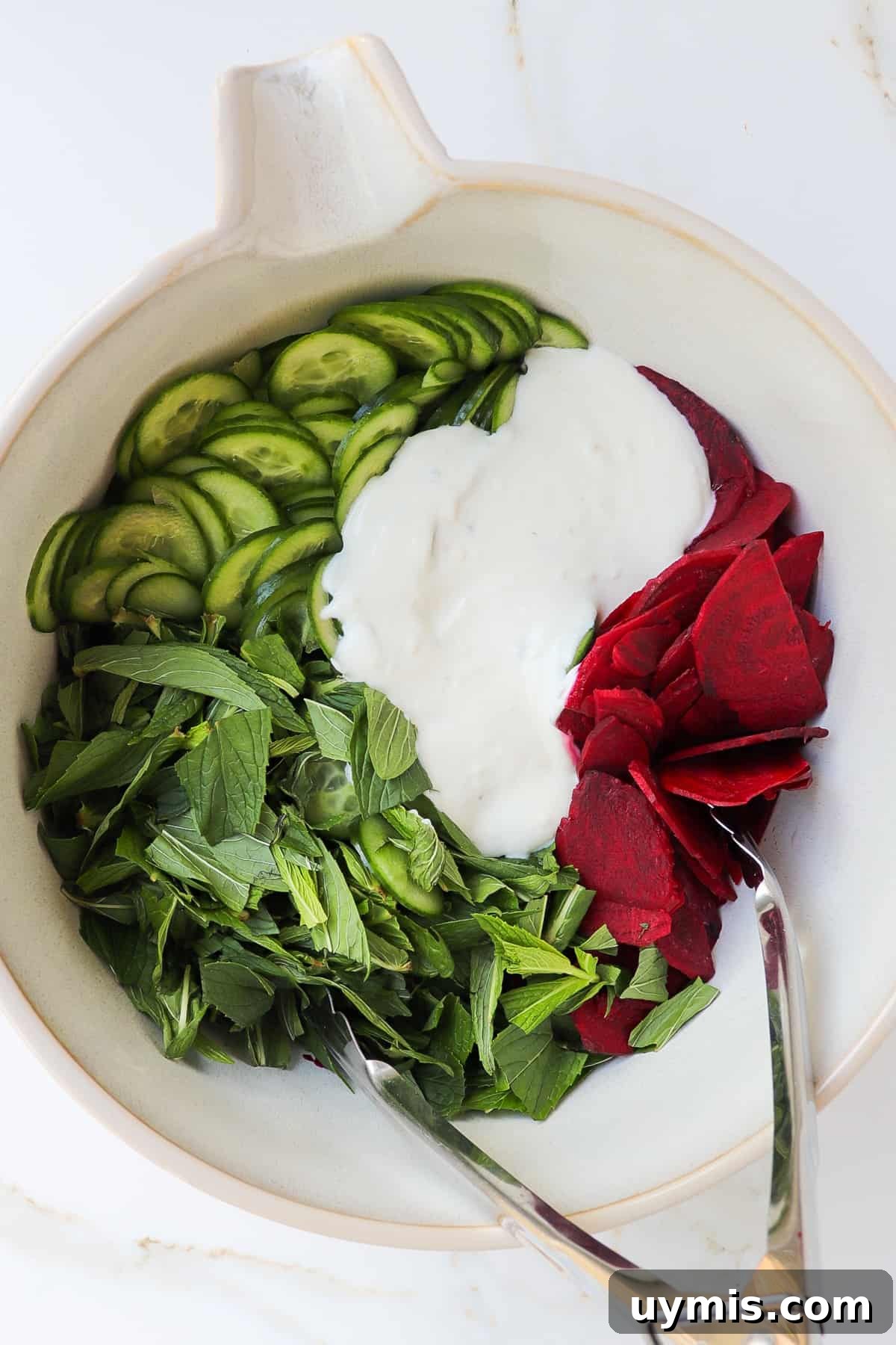 Cucumber and Beetroot Salad Recipe 6 Sliced cucumbers, beetroots, mint leaves and yoghurt dressing in a mixing bowl with metal tongs.