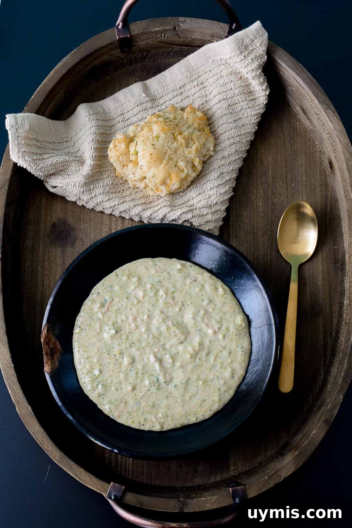 Creamy Homemade Broccoli Cheddar Soup Recipe 6 Soup served in black bowl on wooden platter with gold spoon and biscuit overhead shot