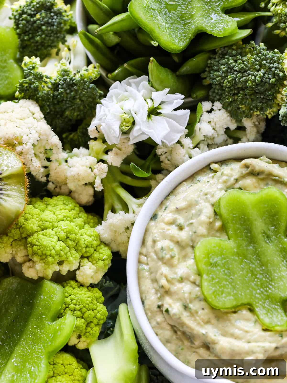 St. Patrick's Day Green Snack Ideas for Parties 5 A small white bowl filled with green dip garnished with a clover shape cut out of a green bell pepper with different green fruits and vegetables.