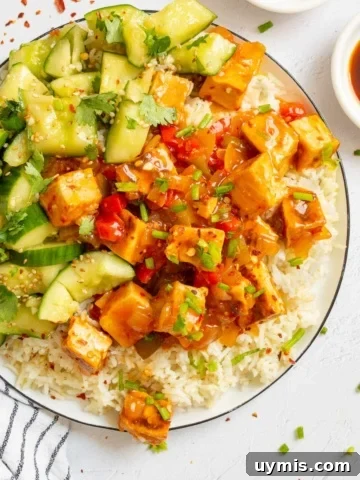 A white plate with tofu in a sticky orange sauce on top of rice with a side of cucumbers.