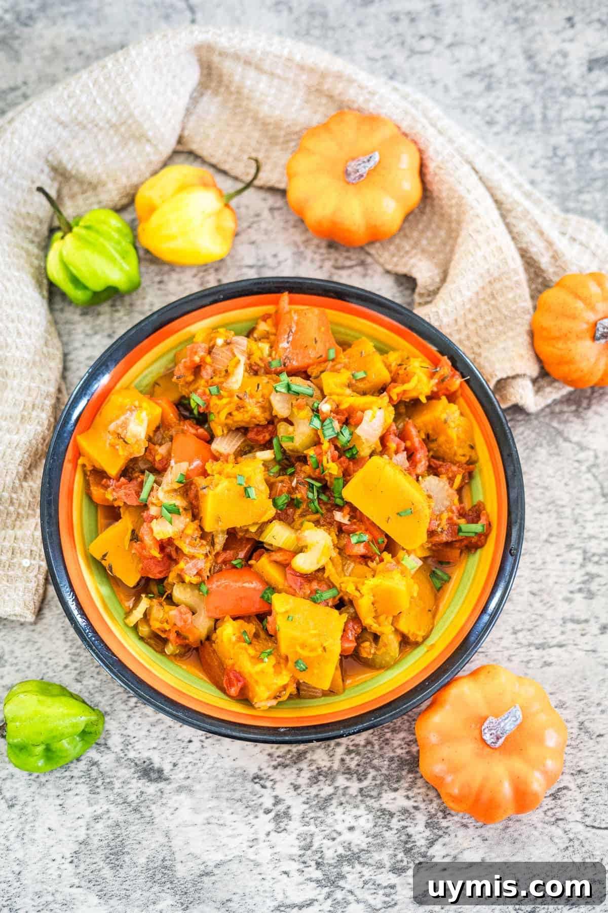 A bowl of cooked mixed vegetables sits on a textured surface, garnished with chopped herbs. Small ornamental pumpkins and peppers are placed around the bowl, including a vibrant Caribbean Spiced Pumpkin. A beige cloth is draped in the background.