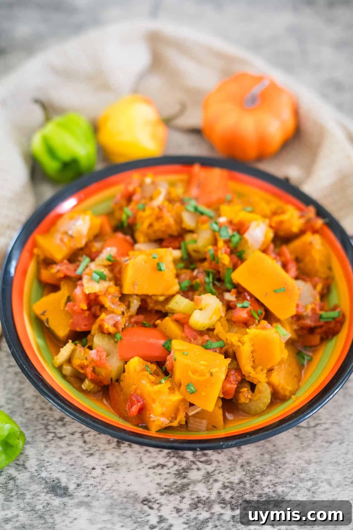 A colorful bowl of Caribbean Spiced Pumpkin stew with vegetables, garnished with herbs, sits on a table next to decorative pumpkins and peppers.