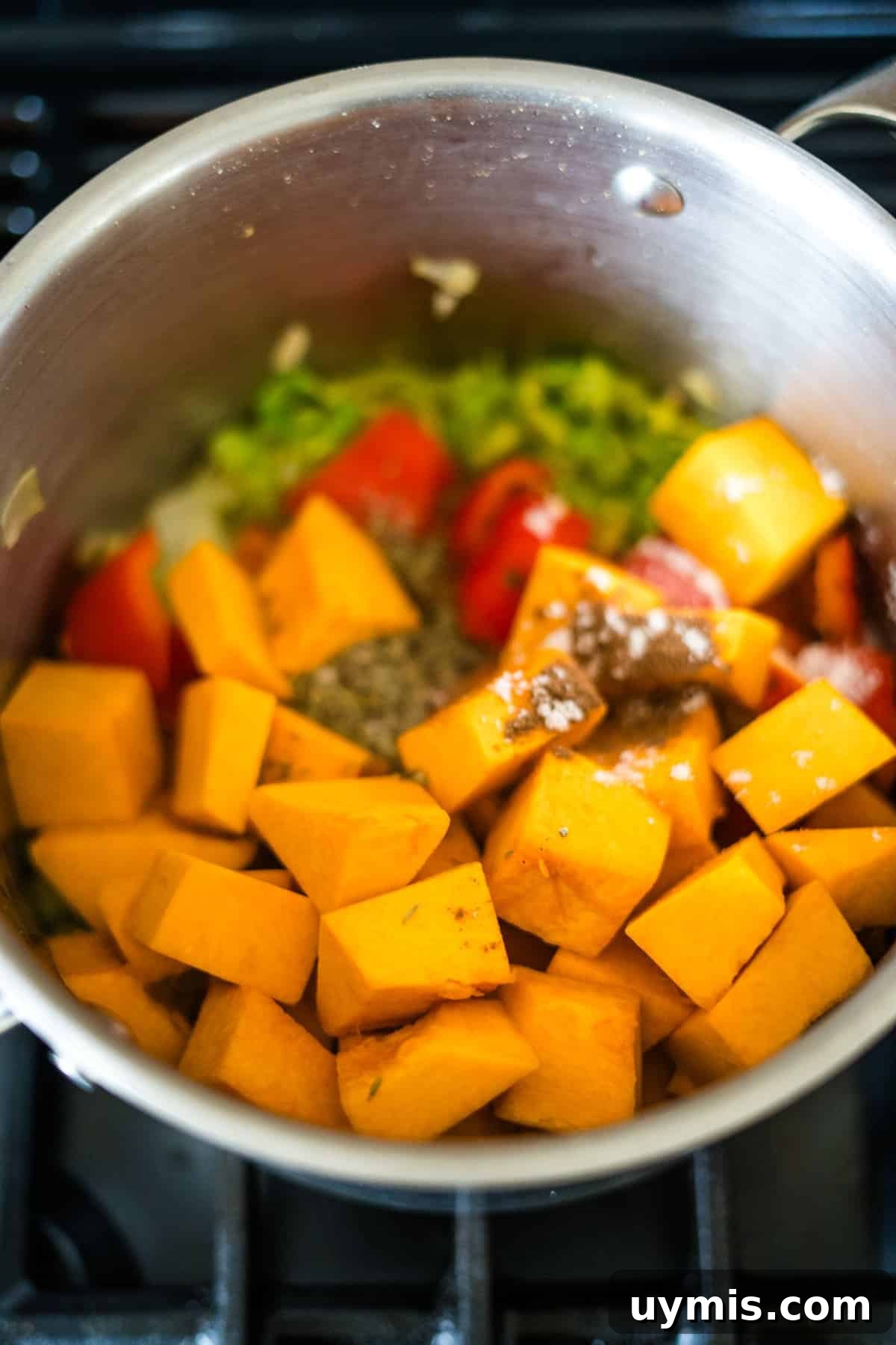 Stainless steel pot on a stove containing chopped butternut squash, bell peppers, Caribbean spiced pumpkin seasonings, and other ingredients being cooked.