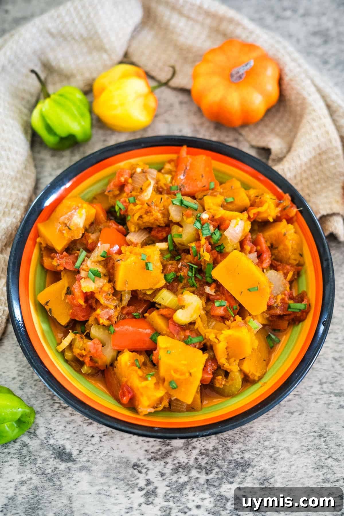 A colorful bowl of cooked vegetables including squash, carrots, tomatoes, and onions, garnished with chopped herbs. Caribbean spiced pumpkin adds a unique twist. Small decorative gourds are in the background.