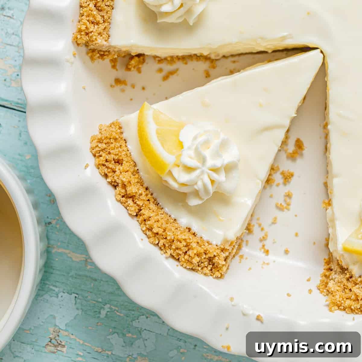 Lemon Icebox Pie Recipe with Creamy No Bake Filling 2 A lemon cream pie with a graham cracker crust in a white dish, with one slice missing and lemon wedge garnish, next to a cup of coffee on a blue wooden surface.