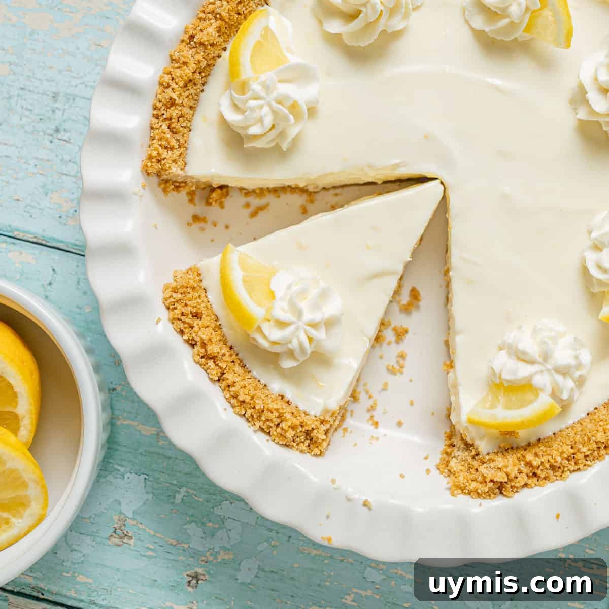 Lemon Icebox Pie Recipe with Creamy No Bake Filling 14 A lemon cream pie with a graham cracker crust, topped with whipped cream and lemon slices, sits on a white plate with one slice removed. A bowl of lemon wedges is beside it.