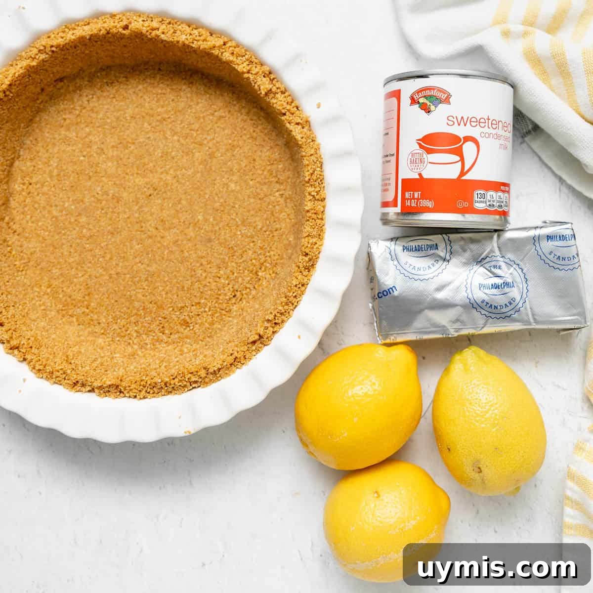 Lemon Icebox Pie Recipe with Creamy No Bake Filling 3 A graham cracker pie crust, a can of sweetened condensed milk, a block of cream cheese, and three lemons on a white surface.