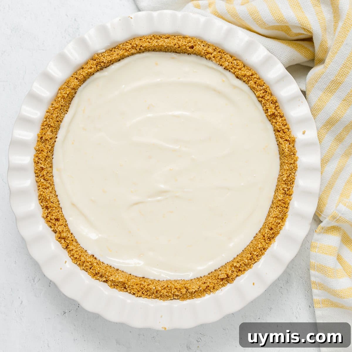Lemon Icebox Pie Recipe with Creamy No Bake Filling 7 A pie dish with a graham cracker crust filled with a smooth, creamy white filling sits next to a yellow and white striped cloth.