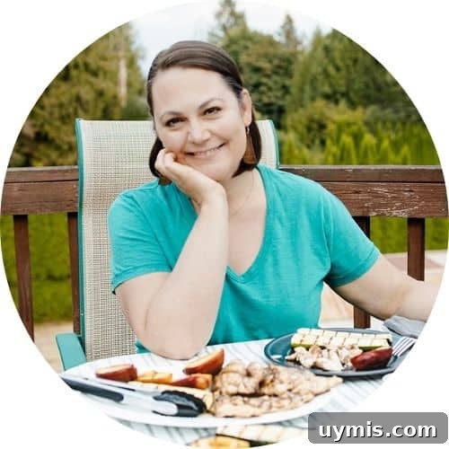 Grill Like a Mother Podcast: Grilling Recipes, Tips & Stories 3 A smiling woman with short dark brown hair in a teal t-shirt sitting outdoors on a deck at a table with grilled food on plates in front of her.
